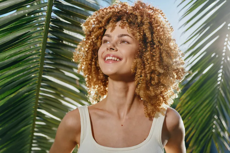 smiling woman with curly hair enjoying tropical sunlight stands near green palm leaves, wearing white tank top, expressing happiness and natural beauty outdoors