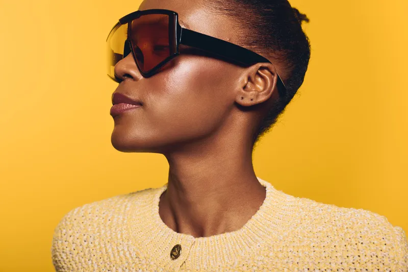 Stylish African woman with dark skin wearing large sunglasses and a yellow knitted sweater poses sideways on a bright yellow background. Concept for fashion, beauty, and summer style.