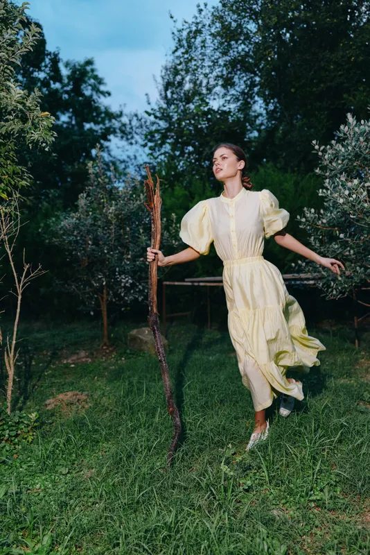 A woman in a pale yellow dress walks outdoors in a lush garden, holding a wooden staff, her gaze forward, capturing a graceful, vintage inspired moment in nature