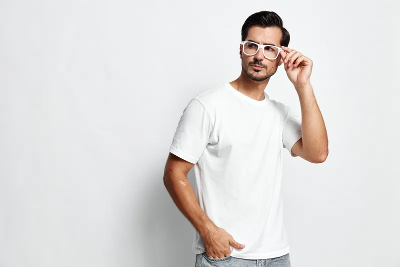 Stylish young man in white t-shirt and trendy glasses looking confident while posing against a plain gray background in an isolated studio portrait. Fashion concept.