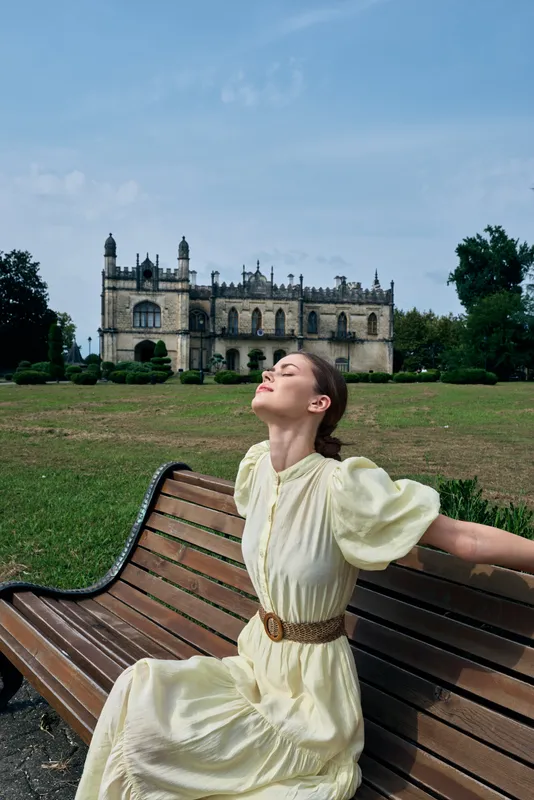 A portrait of a woman in a pale vintage dress sits on a curved bench in a sunny park, eyes closed, head tilted up, enjoying the breeze with a historic castle in the distance.