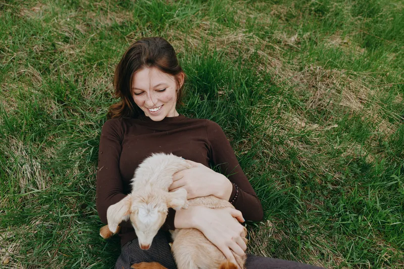 Young woman lies on green grass holding a baby goat in her arms while smiling gently in a relaxed outdoor lifestyle moment.