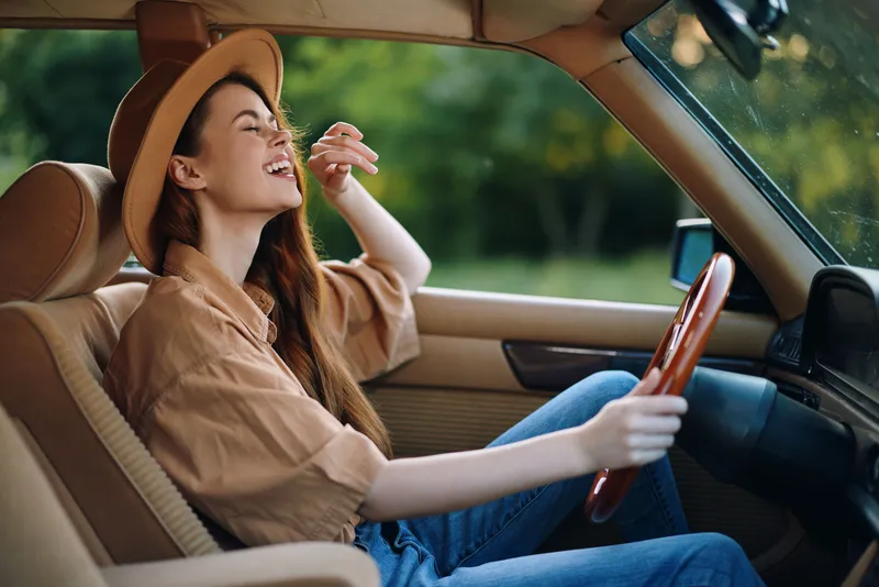 Young woman laughing joyfully in a vintage car, wearing a beige shirt and a stylish hat, capturing the essence of freedom and adventure in nature.