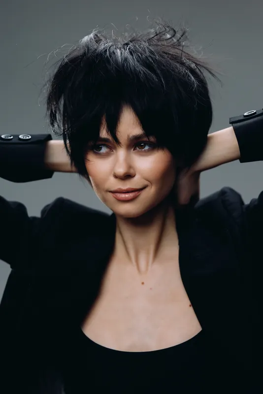 Smiling woman with black short hair wearing a dark outfit, showing confidence and relaxed pose in a studio setting.