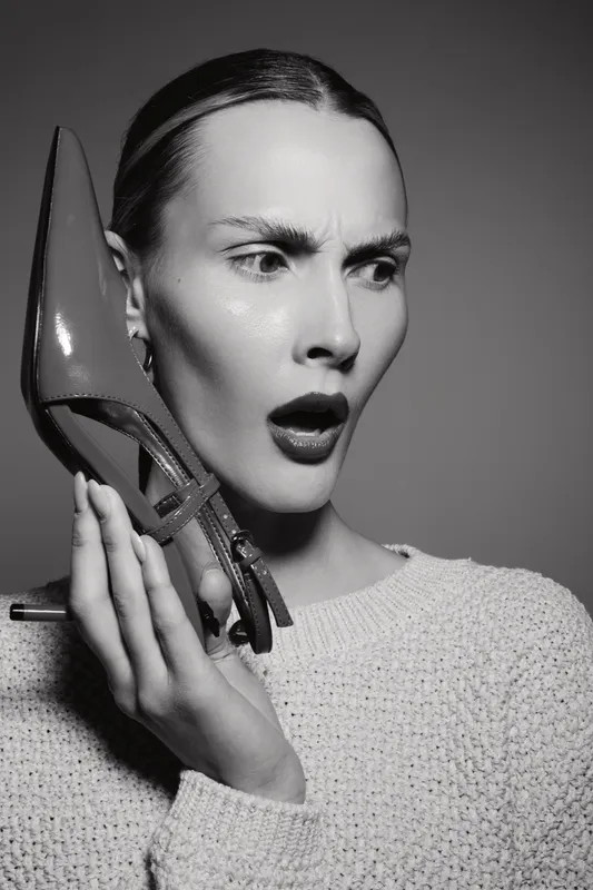 White woman with fair skin and slicked back hair holds a high heel shoe near her ear, expressing surprise or confusion. Black and white close-up portrait with dramatic lighting.