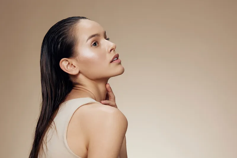 A close up portrait of a young model with long, wet hair and natural makeup, set against a soft beige background, radiating a sense of confidence and beauty