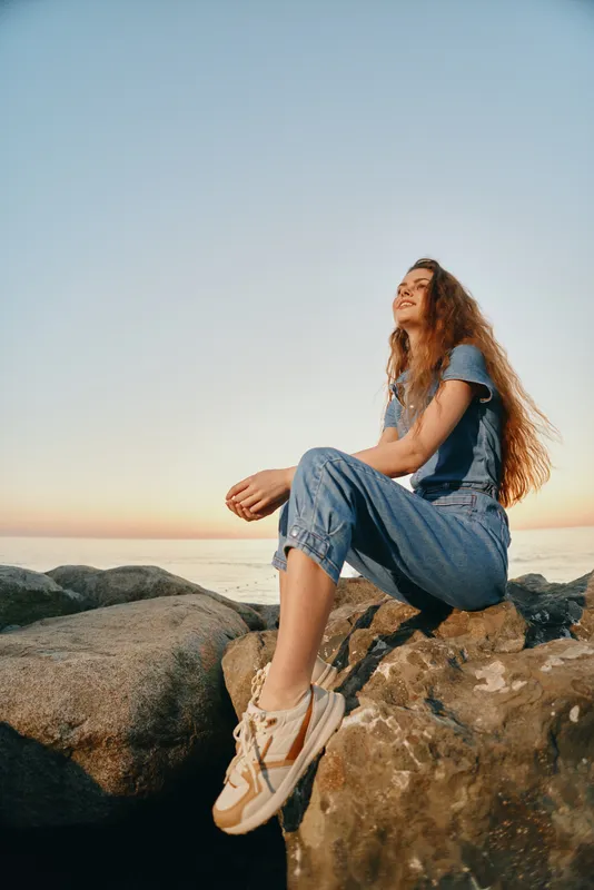 lifestyle woman in modern boho-western denim outfit sitting on rocks by the sea during golden hour film color style