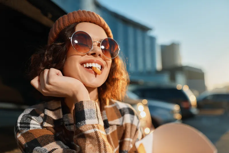 Woman portrait smiling with skateboard in urban parking lot at golden hour, candid lifestyle shot with beanie and sunglasses, authenticity, mindful living and emotional storytelling.