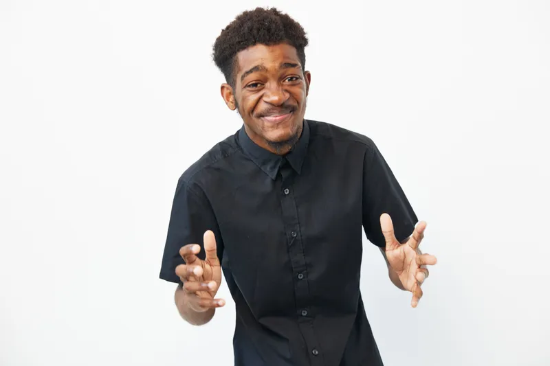 Smiling Black man expressing joy with open arms, wearing a stylish black shirt against a clean white background, radiating positivity and enthusiasm.