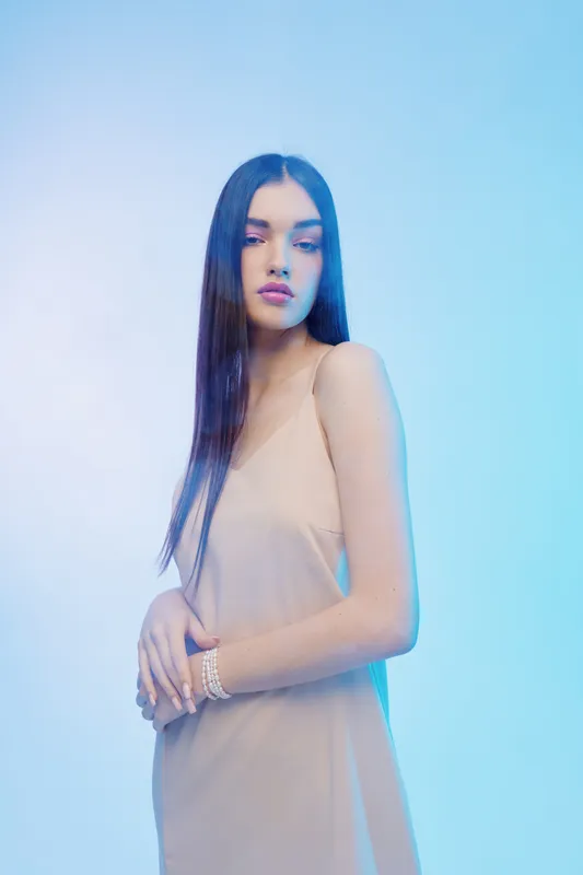 Young woman with long straight hair wearing a beige dress poses with folded hands in soft blue studio lighting. Concept for beauty, fashion, elegance, and style.