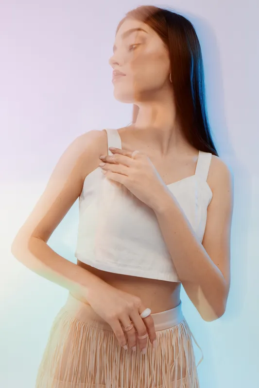 Young woman with long hair poses in a white crop top and beige skirt. Soft lighting creates a dreamy mood. Concept of woman beauty fashion and elegance in studio.