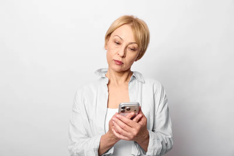Senior woman shows stress and anxiety, holding a smartphone with worry, texting in a quiet studio white background. Mental health concept for communication technology.