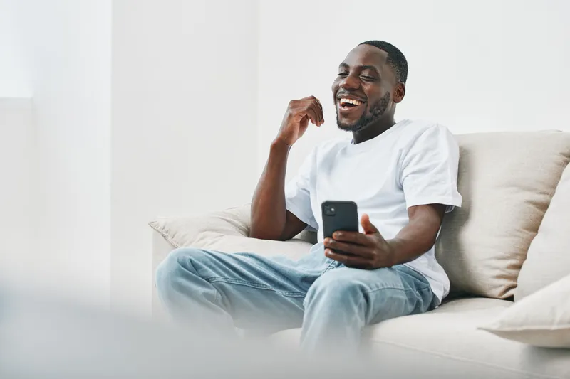 Young African man enjoying time at home, laughing while using smartphone, casual outfit, comfortable indoor setting, bright and cheerful atmosphere