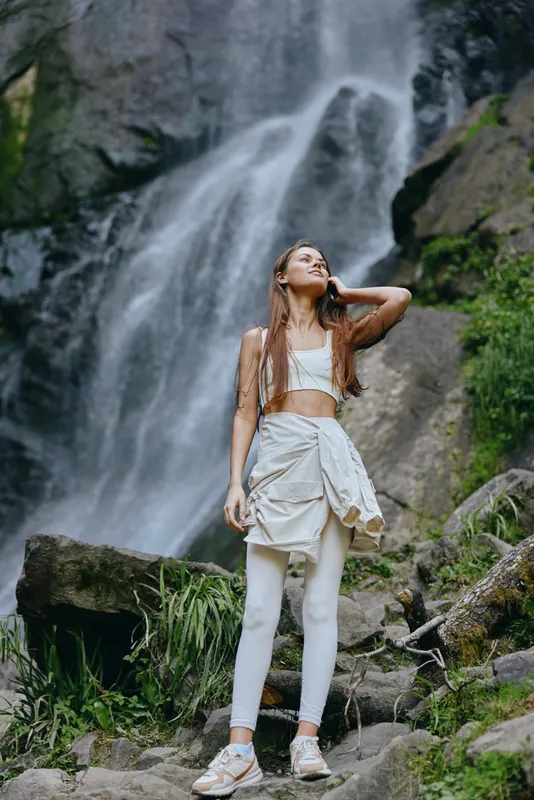 Young woman in athletic wear standing by a waterfall surrounded by rocks and greenery, enjoying nature and outdoor adventure with a calm expression.
