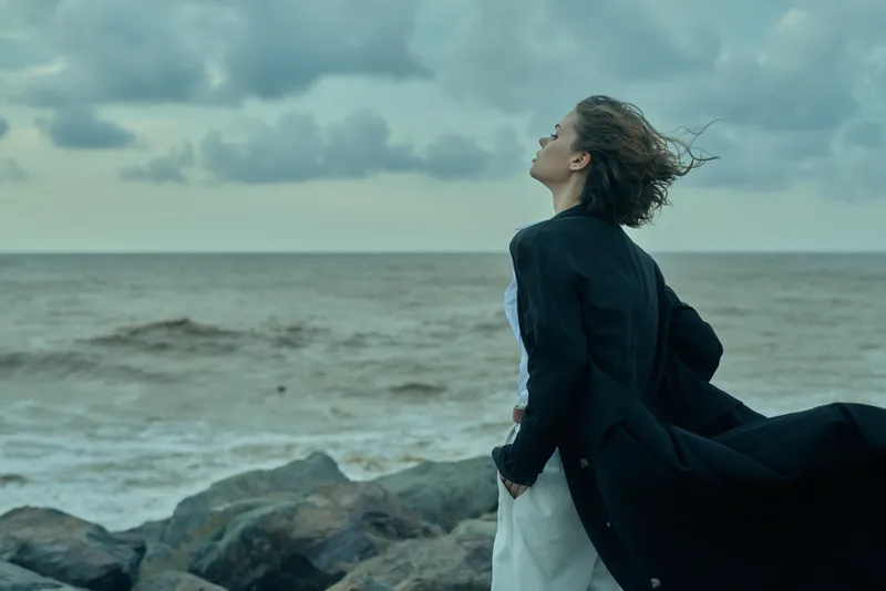 Wind swept dress and coat along the coast by the sea, windblown hair and moody sky, fashion forward silhouette capturing movement and freedom