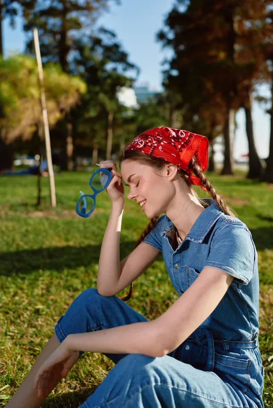 young woman wearing red bandana and denim outfit sitting on grass in sunny park holding blue sunglasses with relaxed expression and casual style