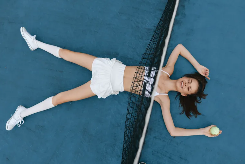 Young Asian woman lying on a tennis court, smiling joyfully, wearing sporty attire and holding a tennis ball, conveying a sense of fun and leisure