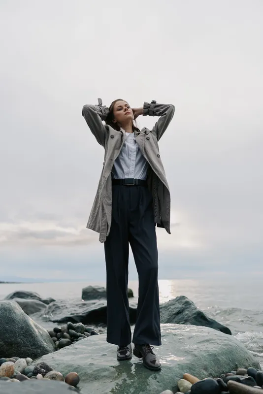 Young woman in stylish coat and wide trousers stands on rocky shore with arms raised behind head. Overcast sky and calm sea create serene mood and fashion concept outdoors.