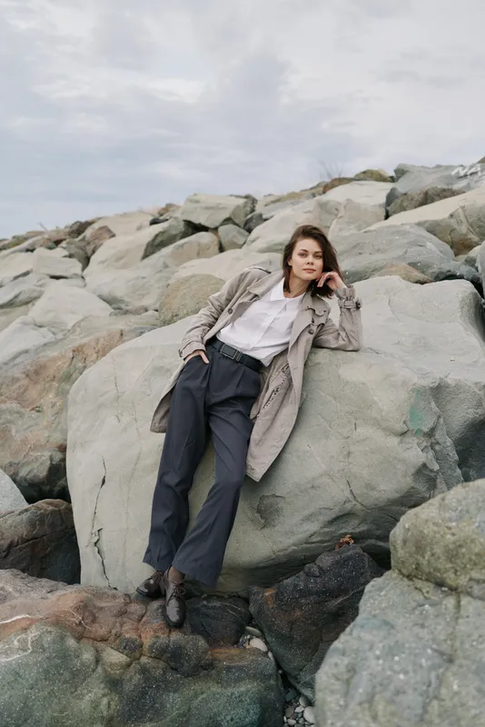 Young woman in stylish outfit leans on large rocks outdoors under cloudy sky. Casual fashion concept with natural background and relaxed pose by the rocky seashore landscape.