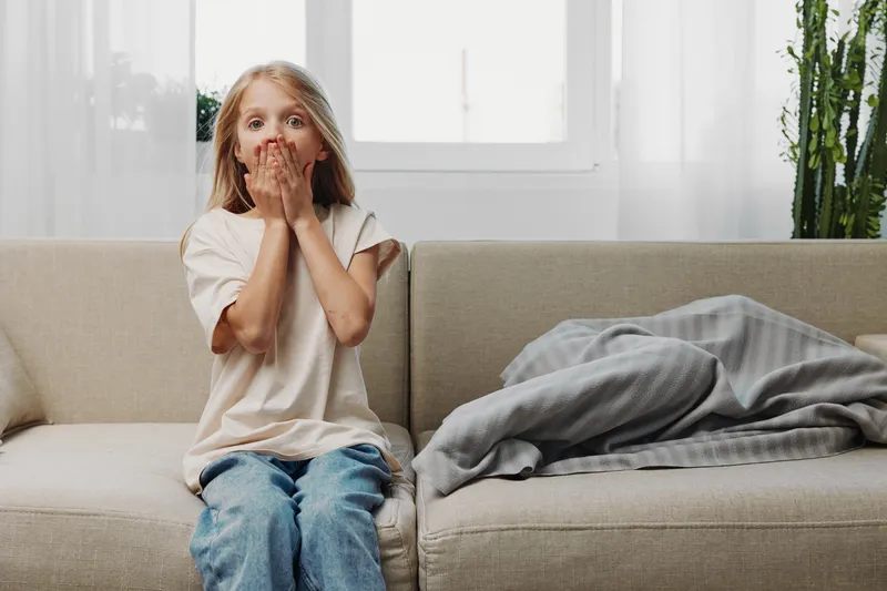 Surprised girl sitting on beige sofa covers mouth with hands in bright living room with large window and plants. Concept of shock and innocence.