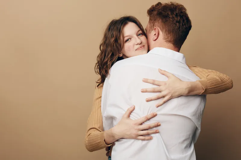 A young couple embraces warmly against a beige background. The woman looks at the camera while hugging the man in a white shirt. Concept of love, affection, and connection.