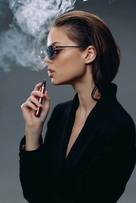 Stylish woman vaping with smoke wearing sunglasses and black jacket indoors lifestyle concept