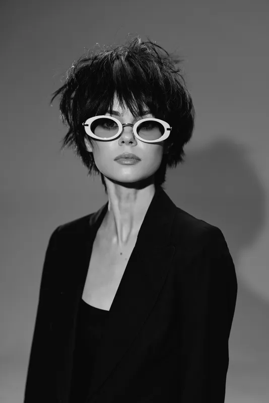 Black and white portrait of a stylish woman with dark hair wearing white sunglasses and a black blazer, expressing confidence and modern fashion.