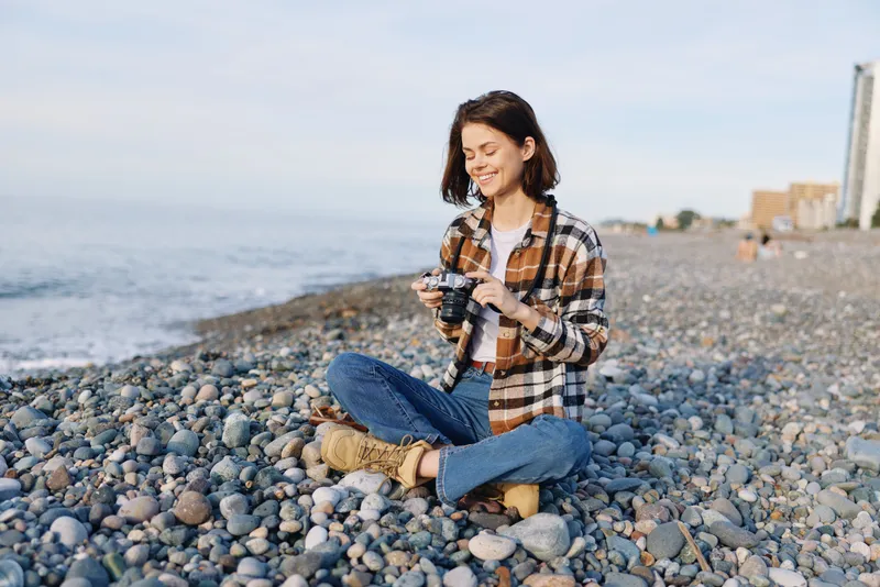 Woman sits on a pebble beach with a camera, wearing a casual plaid shirt and jeans, enjoying the seaside light and a peaceful moment captured in nature.