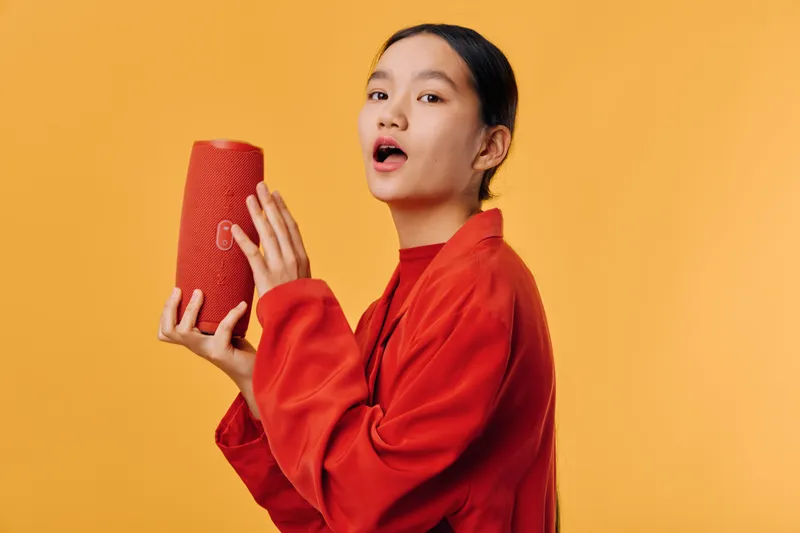 Young woman in red sweater holding portable speaker, singing with open mouth, standing on yellow background. Entertainment, music, technology, and lifestyle concept with happy expression.