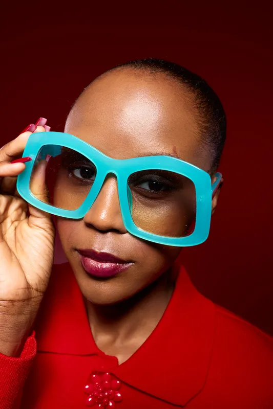 Close-up of a confident dark skinned African woman wearing oversized blue glasses and red clothing against a red background. Concept of fashion, beauty, and bold style.
