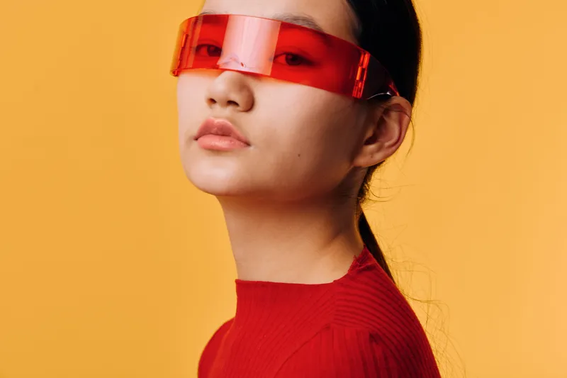 Young woman with short hair wearing futuristic red visor and red sweater against bright orange background. Fashion portrait with modern style and confident look in studio lighting.