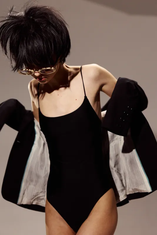 Stylish woman in black bodysuit wearing sunglasses and jacket, posing against a neutral background with confident expression