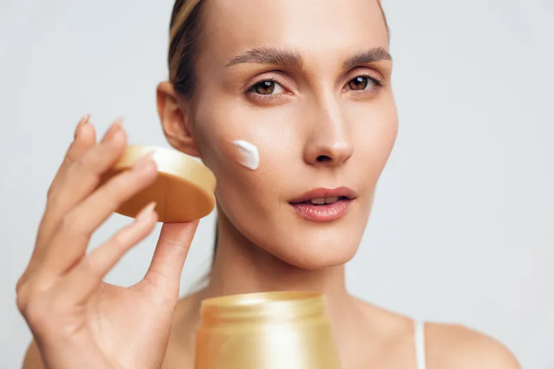 Woman applying white face cream on her cheek with a golden jar in hand. Close-up portrait with natural skin and neutral background. Concept of skincare, beauty, and self-care routine.