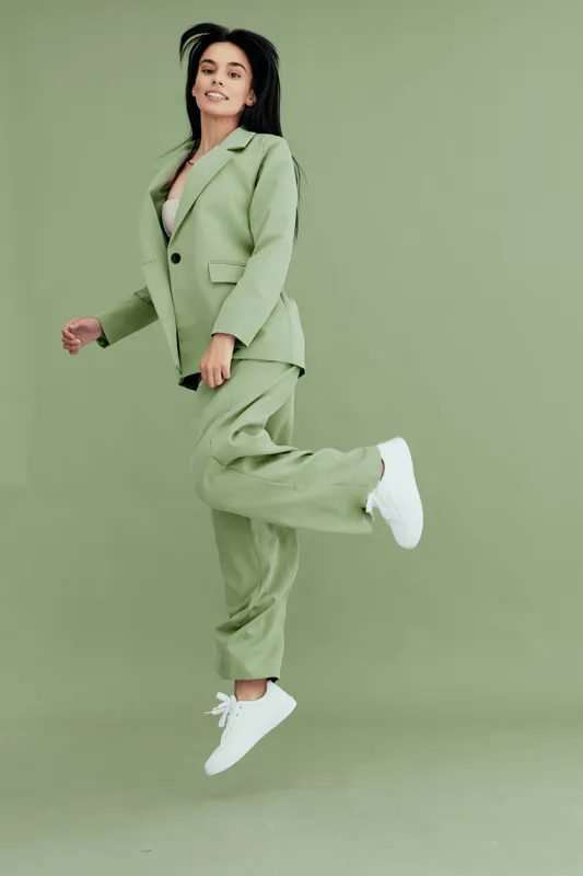 Young woman jumping happily in green pantsuit and white sneakers, smiling confidently against a seamless green background, showcasing joyful motion and stylish casual fashion.