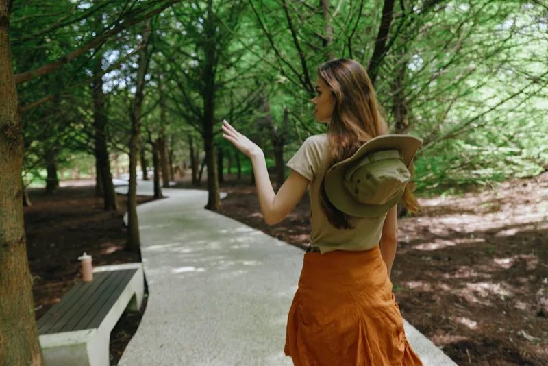 Woman walking with hat in park on tree lined path, casual traveler pausing near a bench, summer fashion and outdoor leisure, back view in lush nature with dappled sunlight.