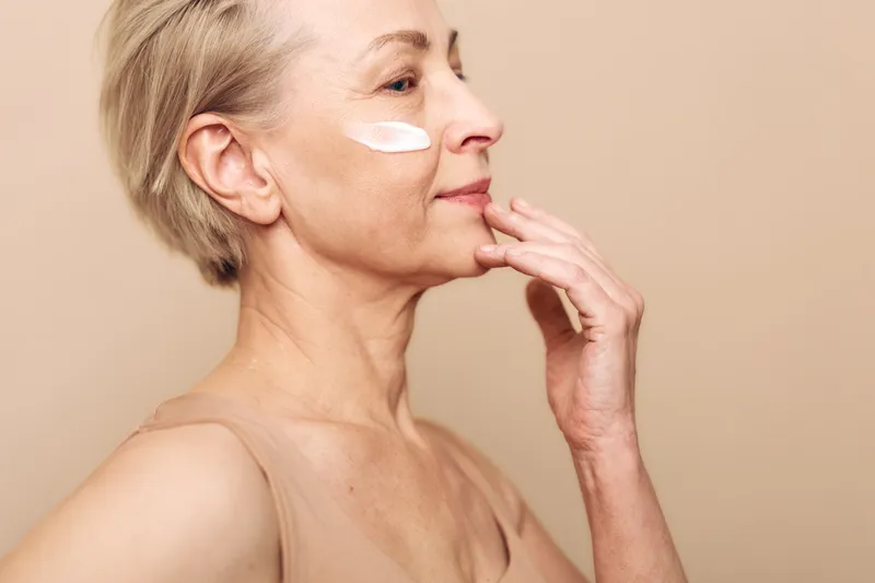 Mature woman with short hair applies white face cream on cheek in a soft beige studio. Concept of skincare, aging skin care, beauty routine, and self-care for older women.