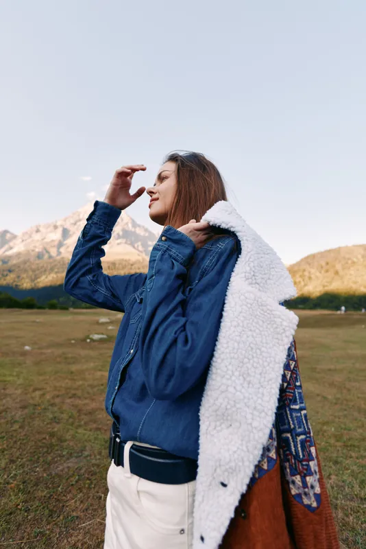 Woman wearing a jacket in mountains outdoors on a grassy field, portrait side view with smile and sunglasses, casual outerwear, autumn light, scenic travel and relaxed vibe.