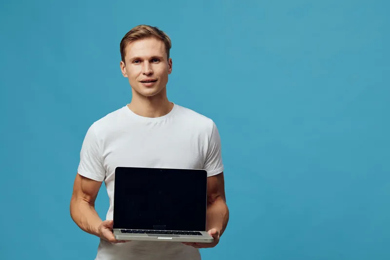Confident young man with fair hair, wearing casual white t-shirt, holds a closed laptop computer facing camera, standing against bright blue background in studio shot, professional technology concept.