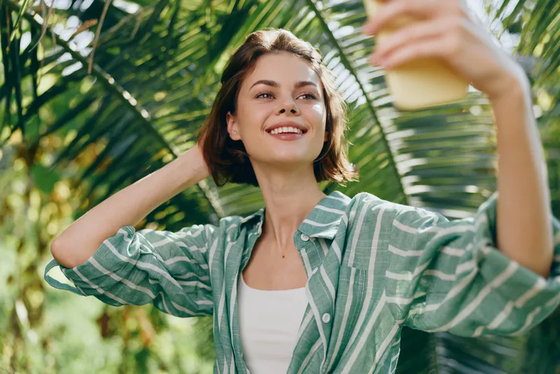Woman outdoors smiling while taking a selfie under bright sun, wearing a striped casual shirt, joyful mood, lush green background, carefree moment, bright lifestyle portrait, sunny day vibe.