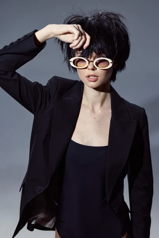 Stylish woman wearing sunglasses and black blazer posing with short hair against gray background