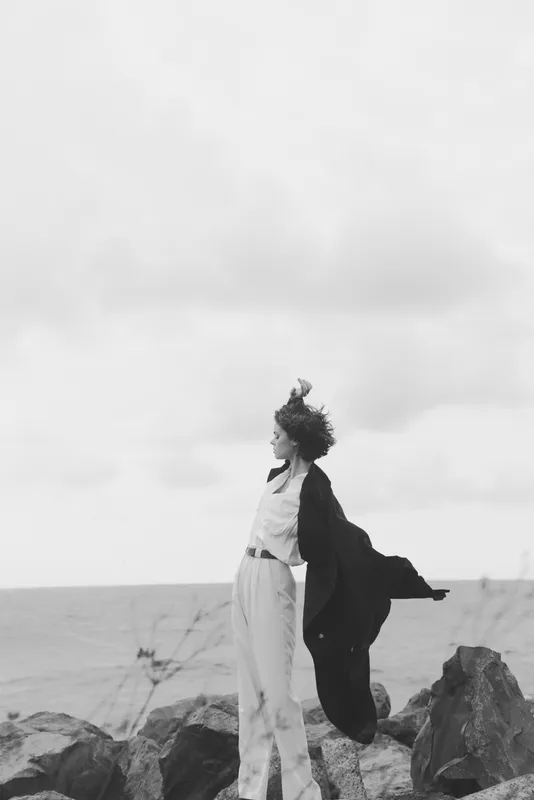 Woman stands on rocky shore as wind lifts a flowing coat, sea horizon behind, calm mood, motion and solitude, outdoor scenery, fashion inspired pose, dramatic lighting, ocean breeze, natural