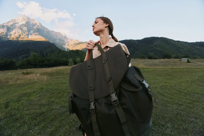 A young woman hiker with a large backpack stands in a wide meadow, mountains in the background, conveying determination, anticipation, and a spirit of adventure.