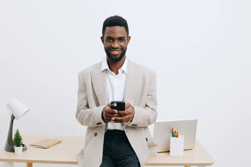 Smiling man standing in modern office with white wall, holding smartphone, casual beige blazer and white shirt, professional businessman using mobile device, lifestyle concept, full body shot, confide