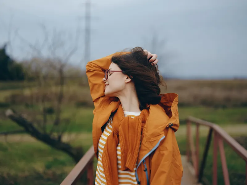 Woman lifestyle outdoors portrait in autumn, orange jacket and glasses, adjusting hair near a bridge, cloudy sky over countryside, calm mood, everyday fashion, with soft wind and trees