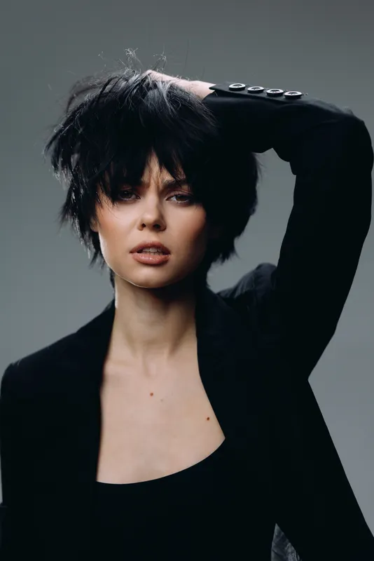 Portrait of a young woman with tousled hair wearing a black jacket and expressive facial expression in a studio setting.