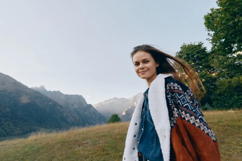 Woman smile in mountains outdoors and nature portrait on a meadow wearing a warm jacket, young traveler enjoying scenic landscape with hair blowing in breeze and peaceful expression.