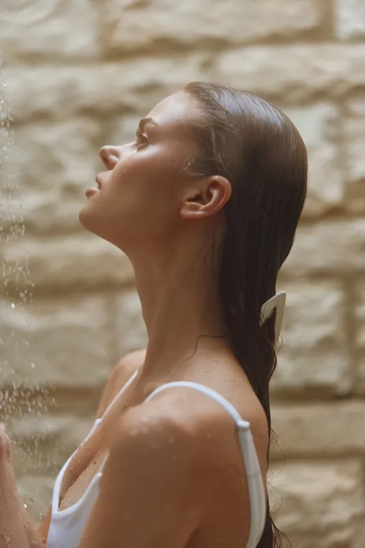 Woman under shower water enjoying a refreshing moment, wet hair, serene expression, natural light, soft background texture, wellness and relaxation theme.