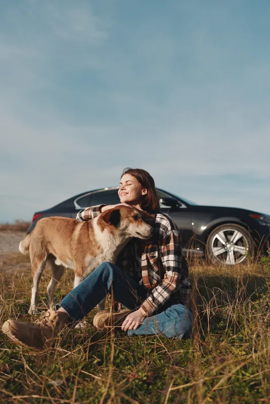 Woman sits on grass with a dog beside her, enjoying a sunny day outdoors, with a car in the background, capturing a calm moment of companionship and nature.