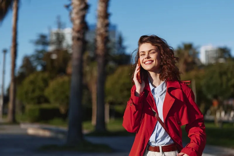 Young woman with curly hair smiles happily while talking on the phone outdoors in a red coat on a sunny day with palm trees in the background.