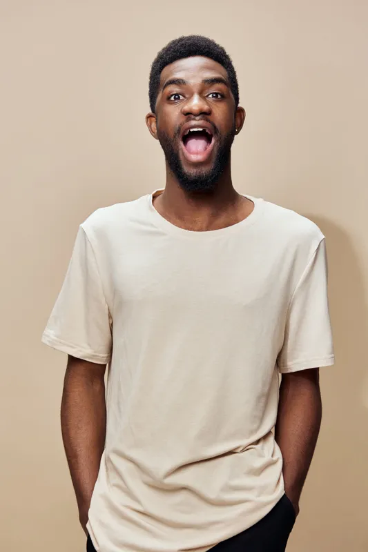 Excited young Black man expressing joy in a light beige t shirt against a neutral background, showcasing happiness, energy, and positive emotions
